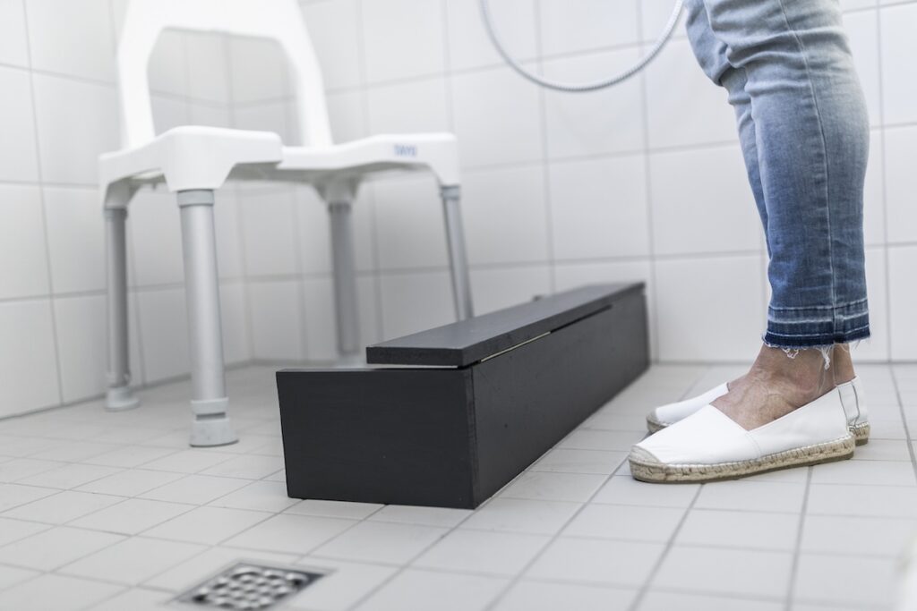 Pflegender steht vor Stuhl in Dusche mit Wasserablauf, um nicht nass zu werden